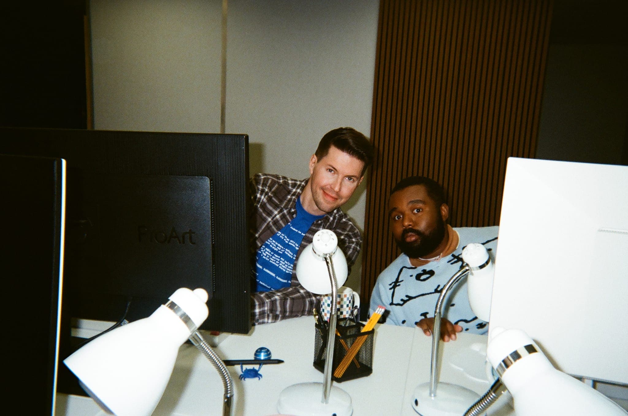 Two crew members peeking over a desk cluttered with monitors and desk lamps